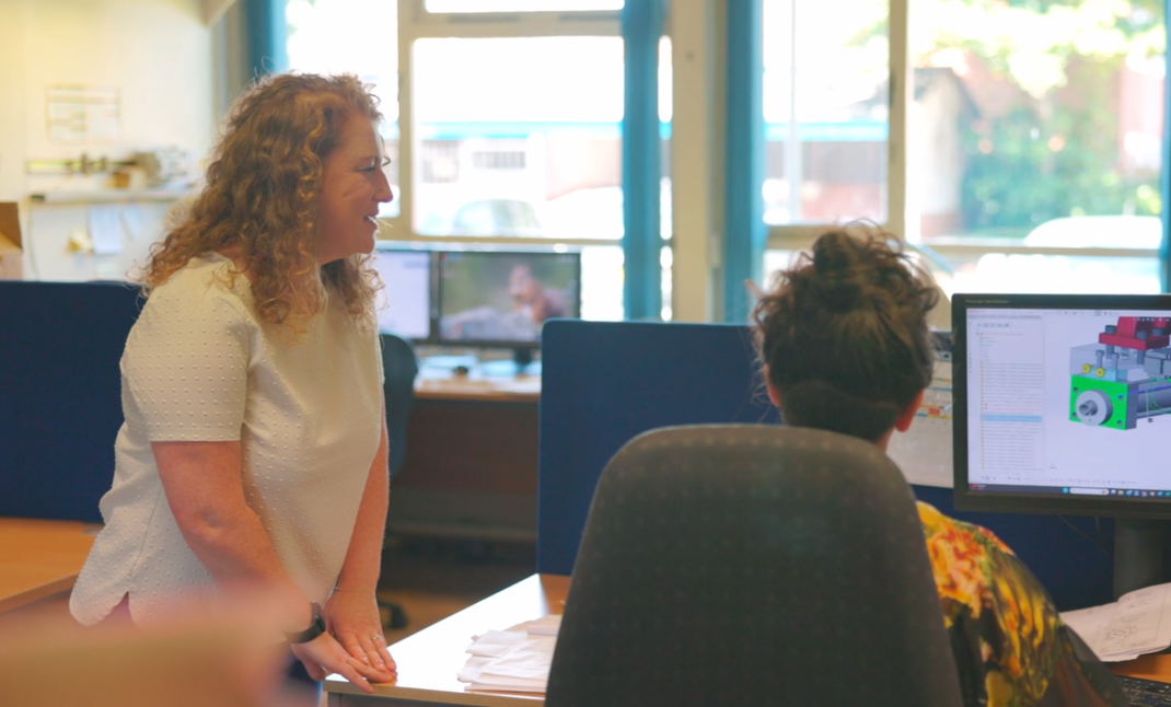 The image shows an office space. A woman sits at a desk with her back to the viewer, looking at an engineering image on a monitor. Next to her, her manager stands and views the screen.